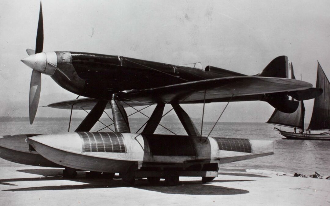 Historic seaplane on beach with sailboat background.