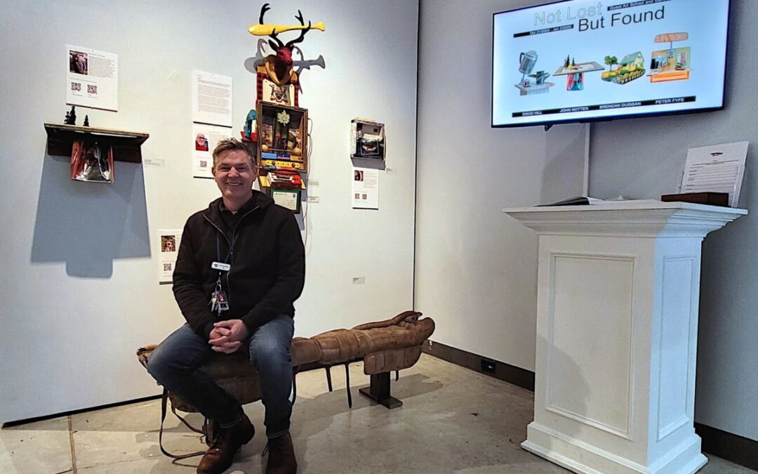 Artist sitting in gallery exhibit room with artwork display.