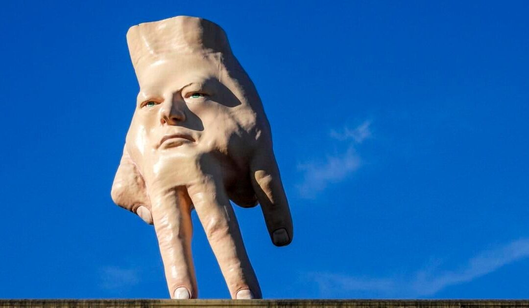 Sculpture of a face-hand hybrid against blue sky.