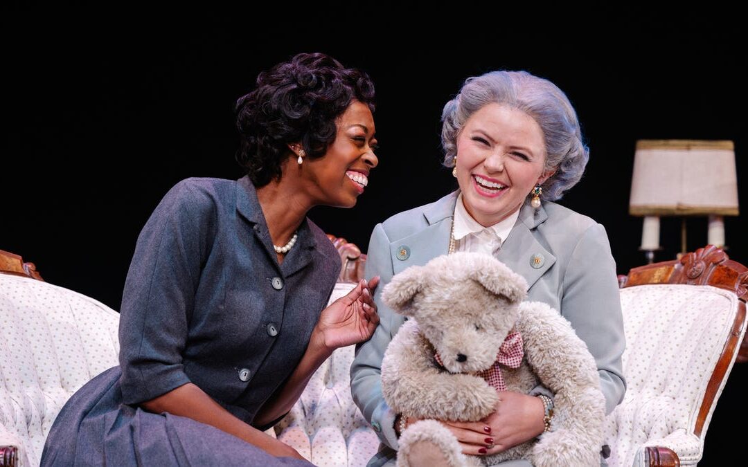 Two women laughing with a teddy bear on stage.