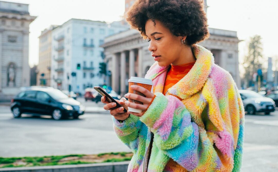 Person using phone, holding coffee, city street background.